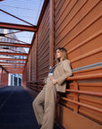 Woman in a beige outfit leaning against a brown wall with a modern architectural background